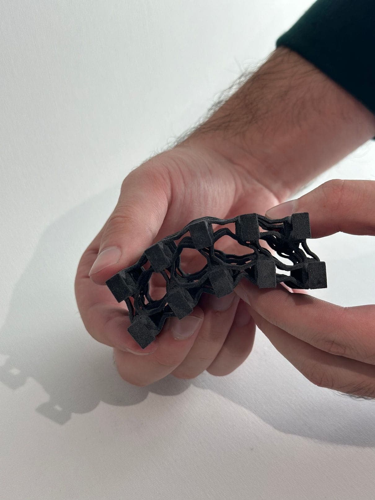 A hand holding a flat sample of the same black lattice against a pale wall. The panel is curving in two directions at once under the pressure of the fingers rather than folding along one line.