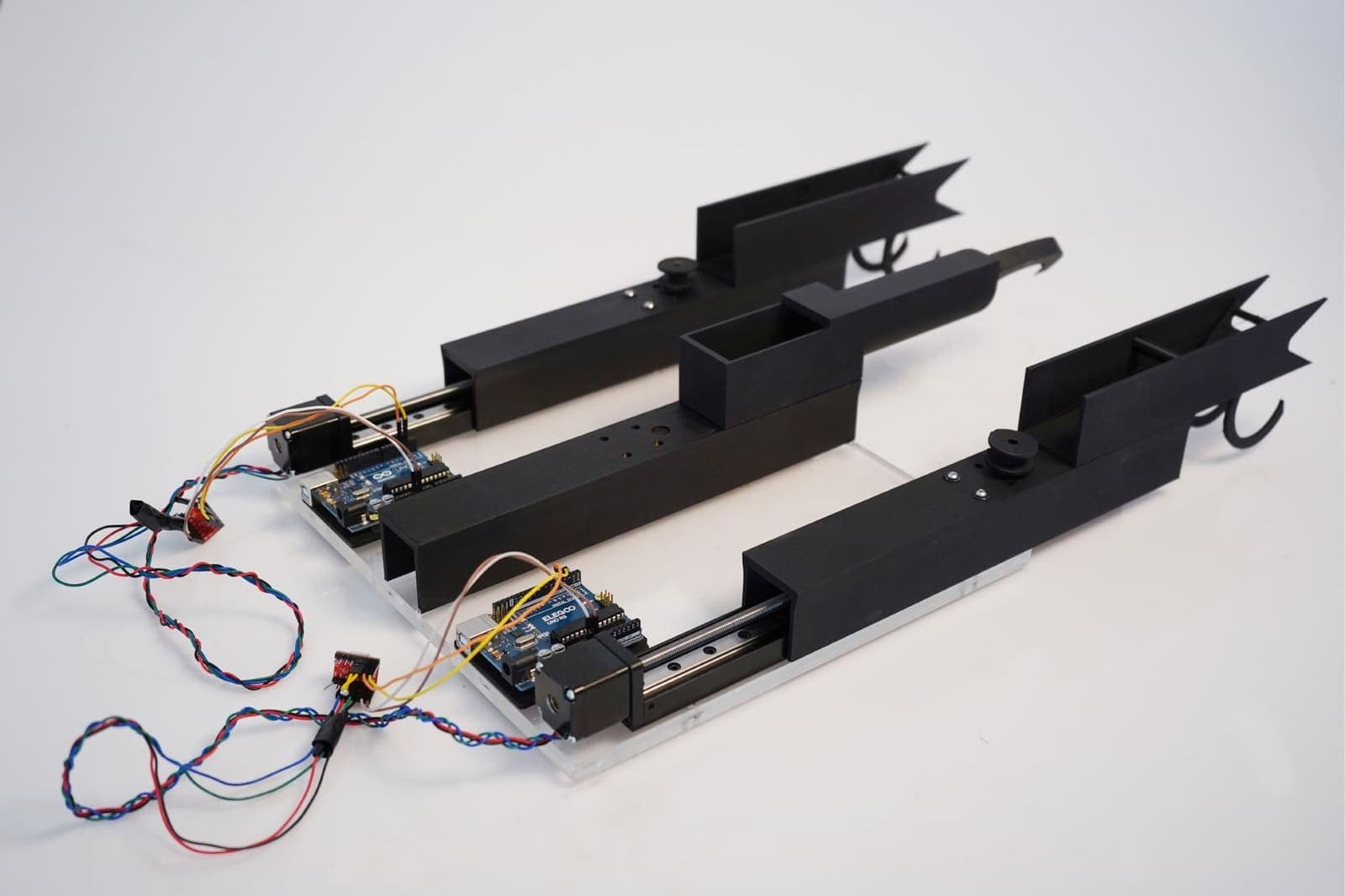 A black prototype photographed on white: two long carriages on linear rails, each ending in a pair of hooked jaws, with stepper motors, exposed control boards and coloured wiring along their length.