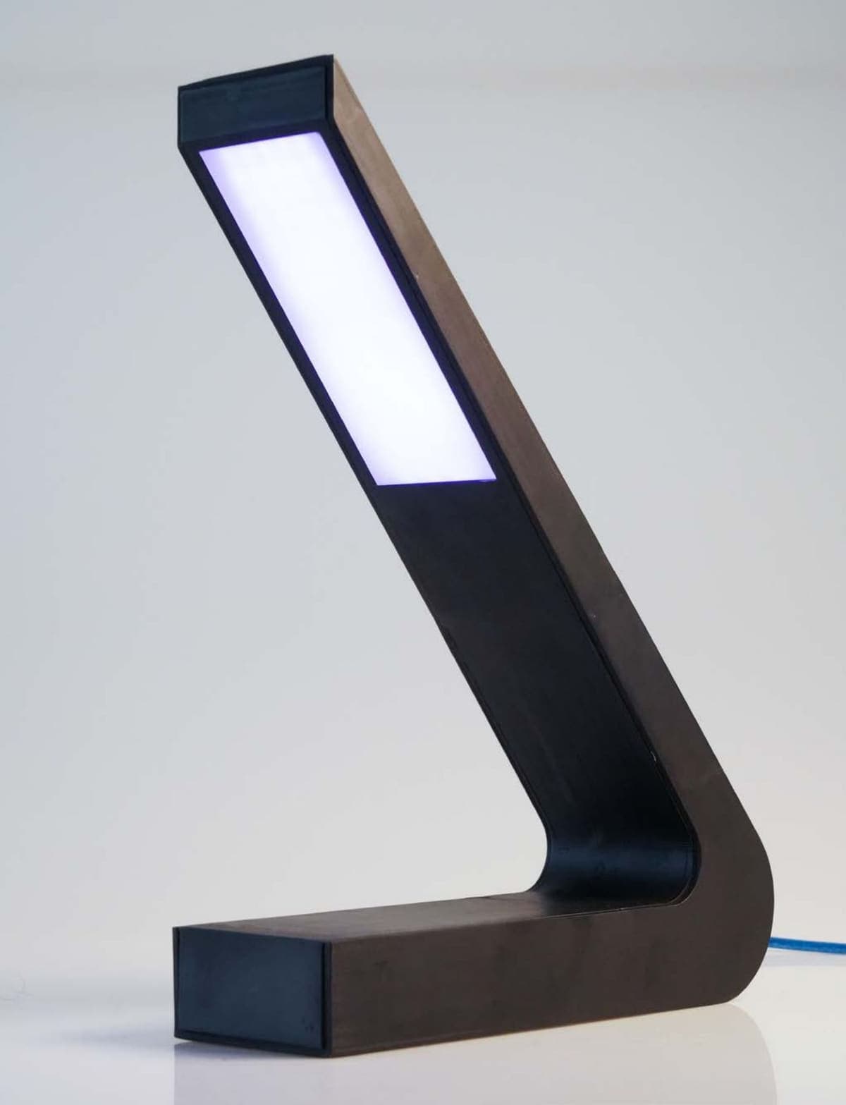 A dark wooden desk lamp shaped like a sundial gnomon — a low foot with an angled arm leaning back from it — the whole upper face of the arm a single lit white panel.